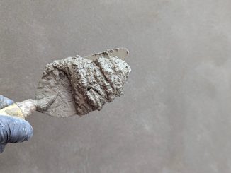 Bàn tay đeo găng cầm bay xây dựng có vữa xi măng, phía sau là mảng tường trát vữa màu xám